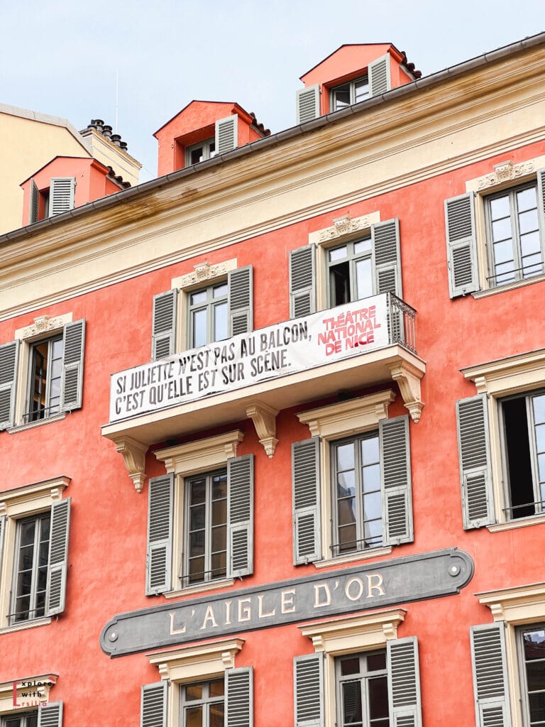 Historic coral-colored building in Nice's Old Town featuring traditional Niçoise architecture with gray shutters, dormer windows, a decorative balcony displaying a Theatre National de Nice banner with a Shakespeare quote in French, and the 'L'Aigle d'Or' (Golden Eagle) sign beneath it, showcasing the city's blend of Italian and French architectural influences.