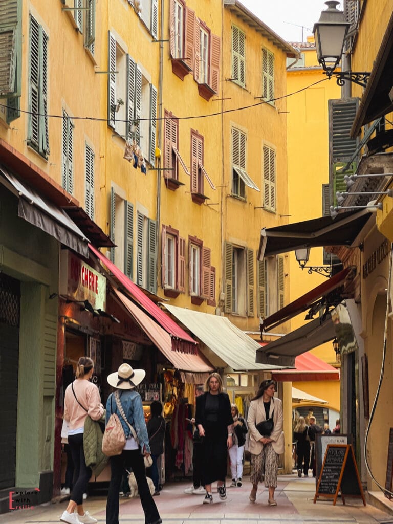 Narrow shopping street in Nice's Old Town featuring traditional yellow Italian-influenced buildings with colorful shutters, canvas awnings over boutique shops, wrought iron street lamps, and tourists exploring the pedestrian alleyways lined with local businesses.