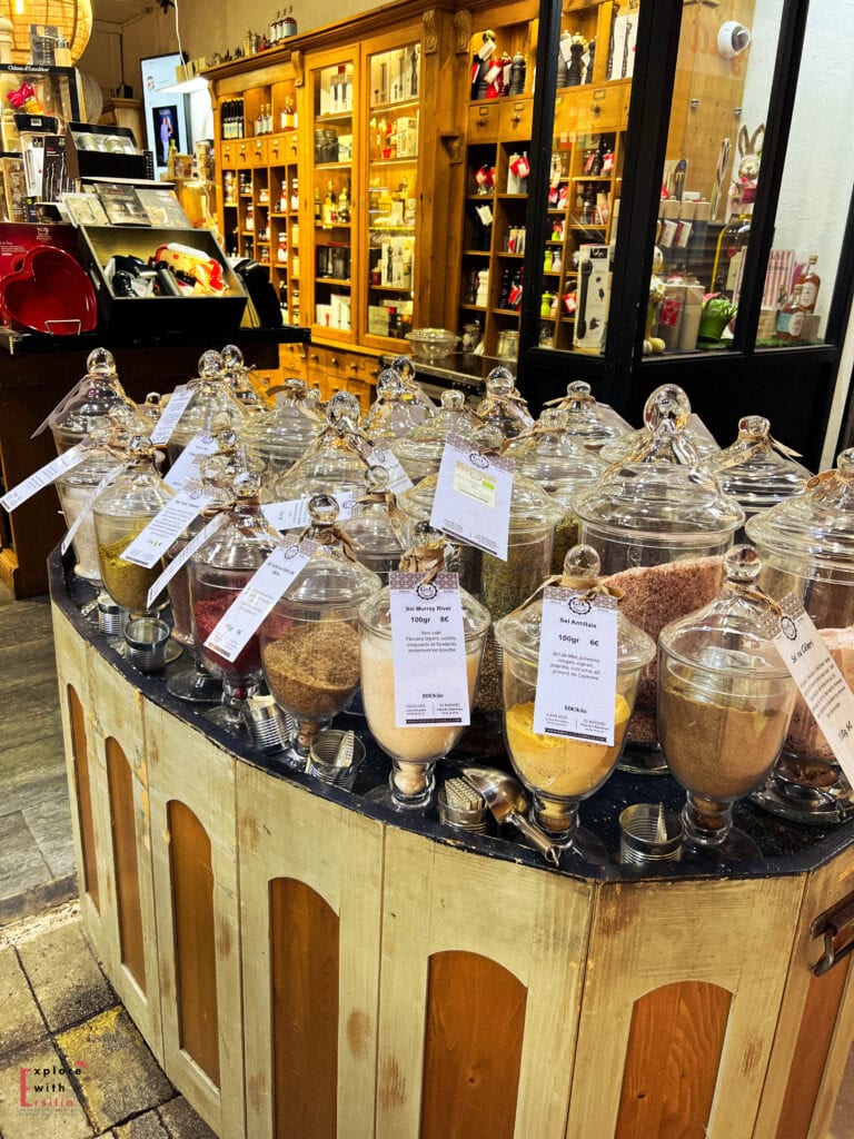 Interior of a traditional spice and specialty food shop in Nice's Old Town featuring glass apothecary jars filled with colorful spices, herbs, and local seasonings on a curved wooden counter, with antique wooden cabinets displaying gourmet products in the background.