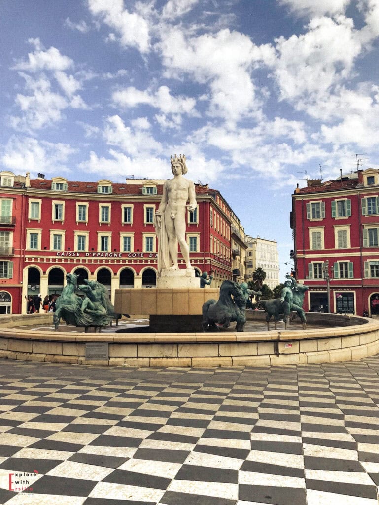 Place Masséna in Nice featuring the iconic Fontaine du Soleil with Apollo statue standing tall in the center surrounded by bronze sculptures, set against vibrant red buildings with white window frames, black and white checkered pavement, and a bright blue Mediterranean sky with fluffy white clouds.