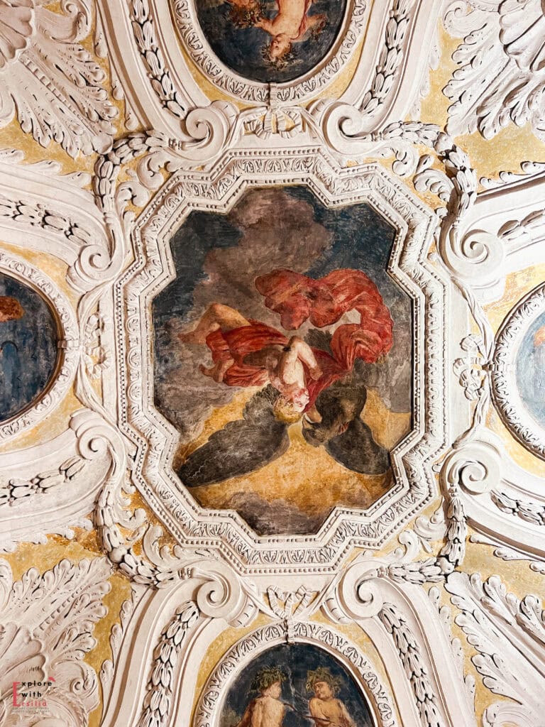 "Ornate ceiling fresco at Palais Lascaris in Nice featuring a mythological scene with figures in flowing red drapery surrounded by elaborate white stucco decorations, gilded accents, and decorative moldings in the Italian Baroque style.