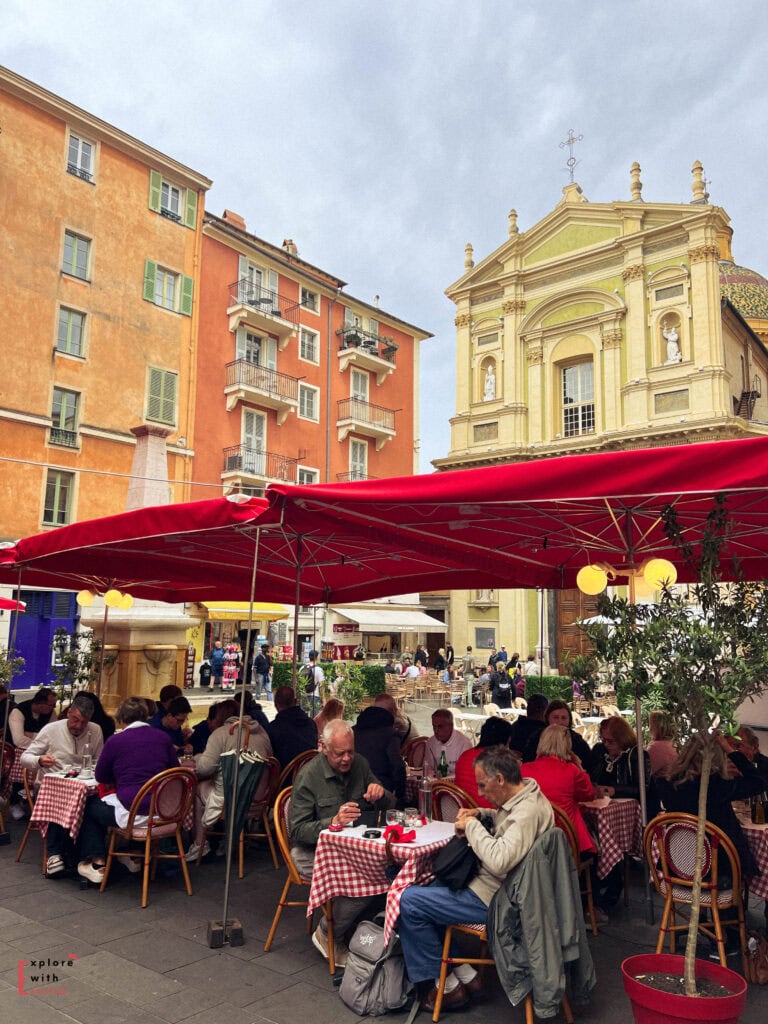 Lively outdoor café scene at Place Rossetti in Nice's Old Town, with diners seated under large red umbrellas at tables with classic red and white checkered tablecloths, surrounded by colorful ochre and orange buildings and the distinctive yellow Baroque façade of Nice Cathedral (Sainte-Réparate) visible in the background.