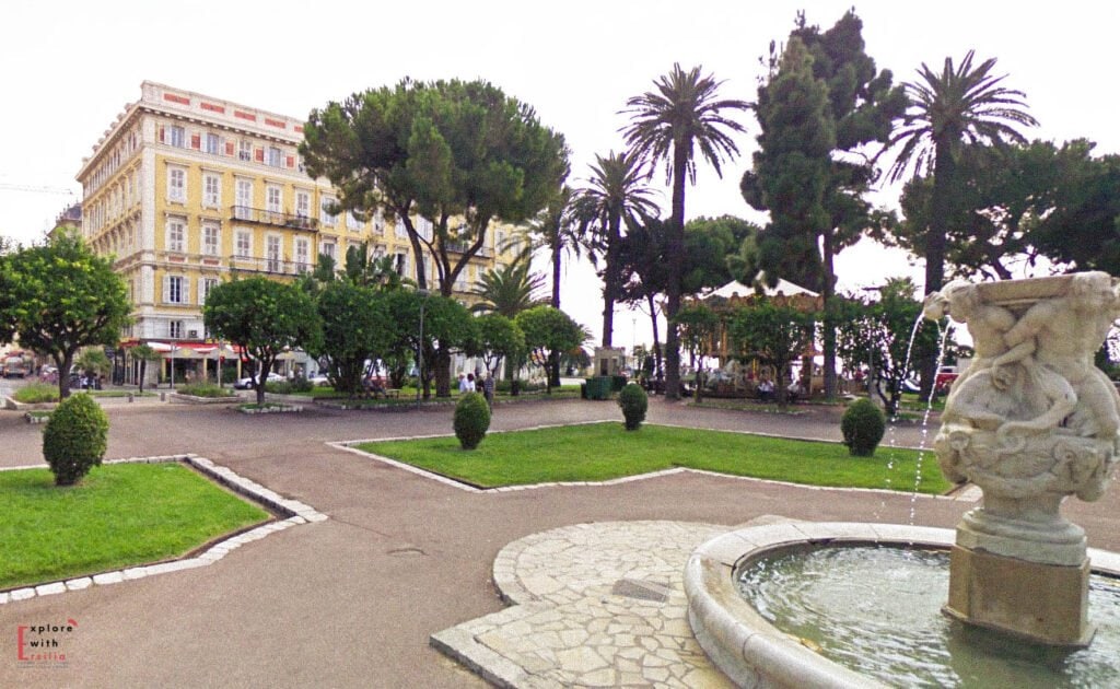 Jardin Albert 1er in Nice featuring manicured lawns, decorative stone fountain with classical sculpture, tall palm trees, umbrella pines, and formal garden pathways, with the elegant yellow Belle Époque architecture of a historic hotel visible across the street.