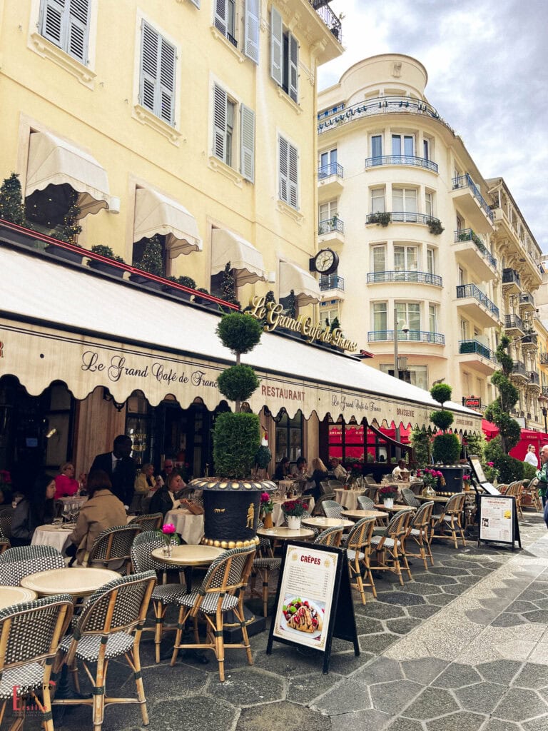 Le Grand Café de France on Rue de France in Nice featuring outdoor terrace with traditional French bistro tables and chairs, cream-colored awnings, elegant façade, and classic rattan seating on stone-paved sidewalks, surrounded by stunning Belle Époque buildings with rounded corner balconies and decorative railings.