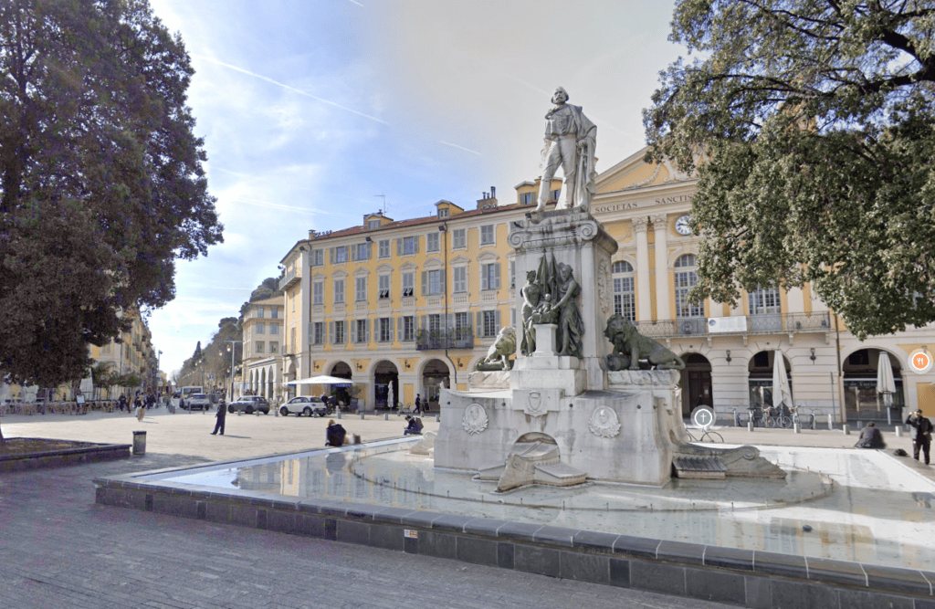 Place Garibaldi in Nice featuring the central Garibaldi monument with marble statues and fountain, surrounded by elegant Baroque-style yellow buildings with arcades, tree-lined edges, and outdoor café seating in this historic square under clear blue skies