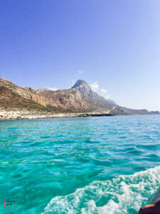 Crystal clear turquoise waters of Balos Lagoon in Crete, viewed from a boat with white wake visible, surrounded by rugged mountainous coastline featuring distinctive peaked formations under a clear blue sky.