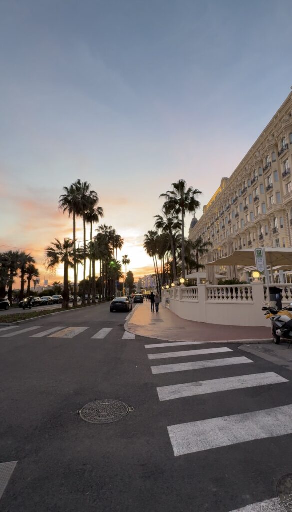 A sunset view of the Cannes waterfront boulevard Promenade de la Croisette, featuring silhouetted palm trees lining the street against a pastel pink and blue dusk sky. The iconic historic white facade of a luxury hotel (possibly the Carlton or Martinez) dominates the right side, with crosswalks and street markings visible in the foreground.