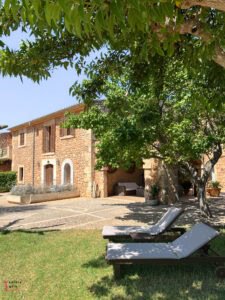 Traditional Mediterranean stone finca with light-colored loungers on a manicured lawn, shaded by lush green trees with an outdoor seating area and terrace under dappled sunlight, showcasing authentic Mallorcan architecture with wooden shutters and natural stone walls under blue sky.