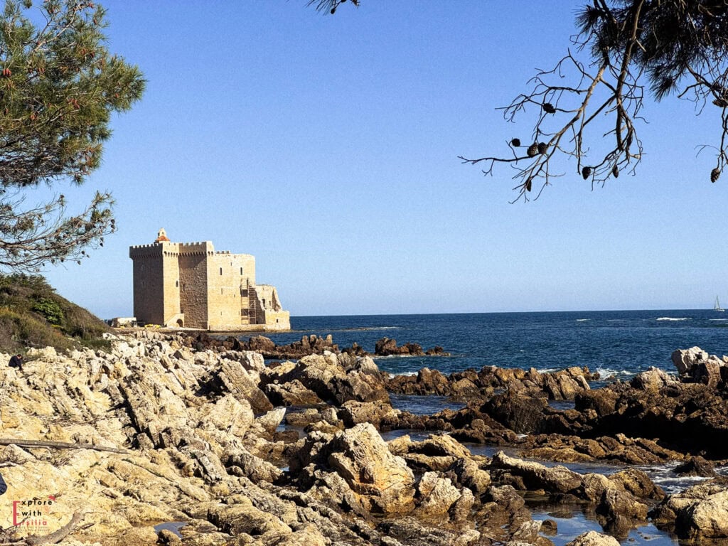 Historic stone fortress on Île Saint-Honorat (Lérins Islands) rising from a rocky coastline against deep blue Mediterranean waters. The medieval square tower fortress stands on the edge of the island, framed by pine trees in the foreground and rugged coastal rocks. Clear blue sky completes this scenic view of the historic monastery island near Cannes.