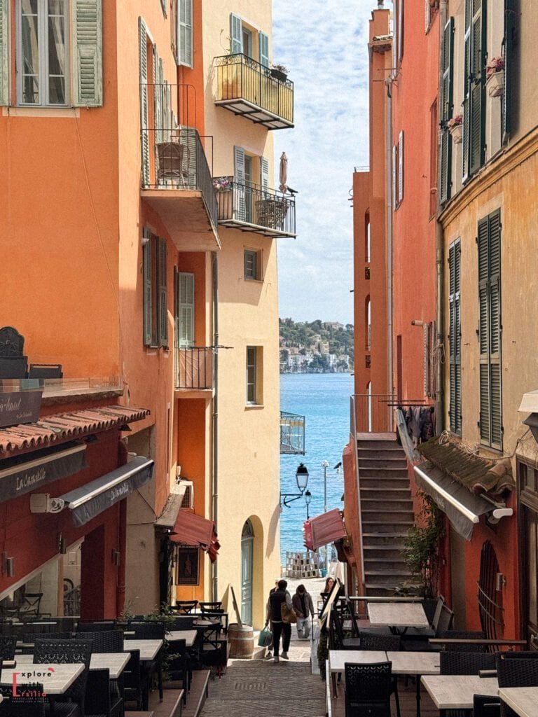 Charming narrow street in Villefranche-sur-Mer with warm-colored buildings in shades of orange, peach, and yellow framing a view of the blue Mediterranean Sea. The picturesque alleyway features traditional shuttered windows, small balconies, outdoor café seating, and stone steps leading down toward the waterfront, creating a quintessential French Riviera scene.