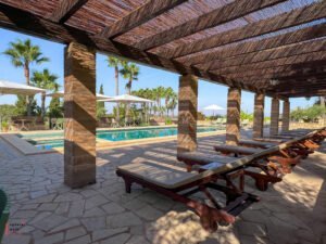 Shaded poolside terrace with traditional reed roof pergola supported by stone columns, featuring wooden sun loungers with cream cushions arranged in a row on stone paving, overlooking a turquoise swimming pool with palm trees and parasols in the background under bright blue Mallorcan sky.