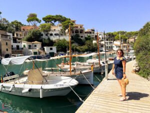 Person in a navy blue dress standing on a wooden dock at Cala Figuera harbor in Mallorca, with traditional wooden boats and fishing vessels moored in turquoise water, surrounded by white Mediterranean houses built on hillsides with pine trees under bright blue sky.