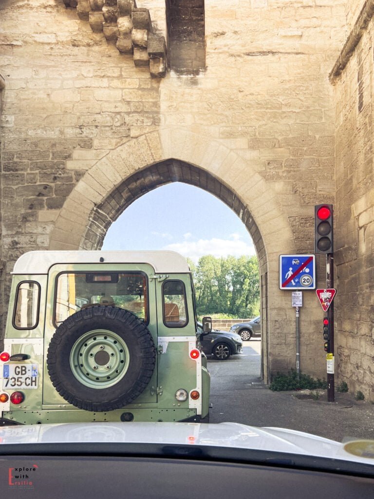 A vintage green Land Rover waits at a red light under a historic stone archway in Avignon, France. The medieval walls frame the narrow passage, blending centuries-old architecture with modern traffic signs.