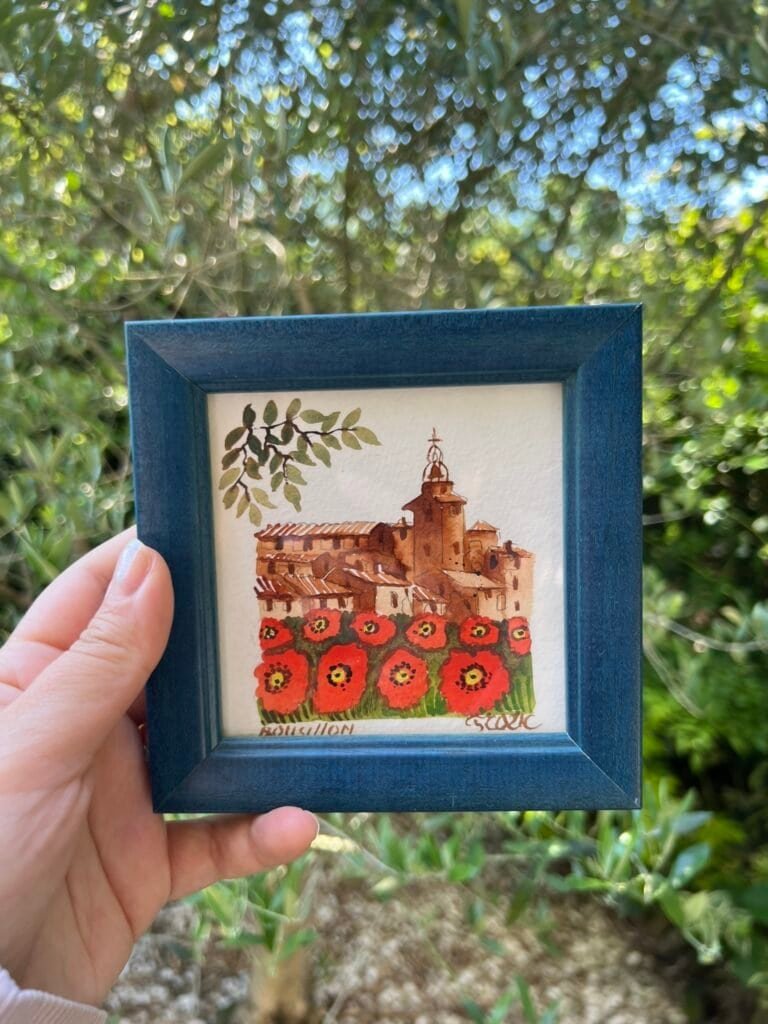 A hand holds a framed watercolor painting of Roussillon, France, showing ochre-toned buildings and a church tower above a vibrant field of red poppies, with leafy green branches in the background.