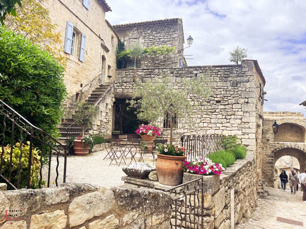 Quaint stone alleyway in Lacoste village, Provence, featuring a charming courtyard with flower-filled pots, café-style tables and chairs, and rustic stone buildings with pastel shutters. The cobbled path leads under an arched gateway, evoking the peaceful, timeless charm of this hilltop village.