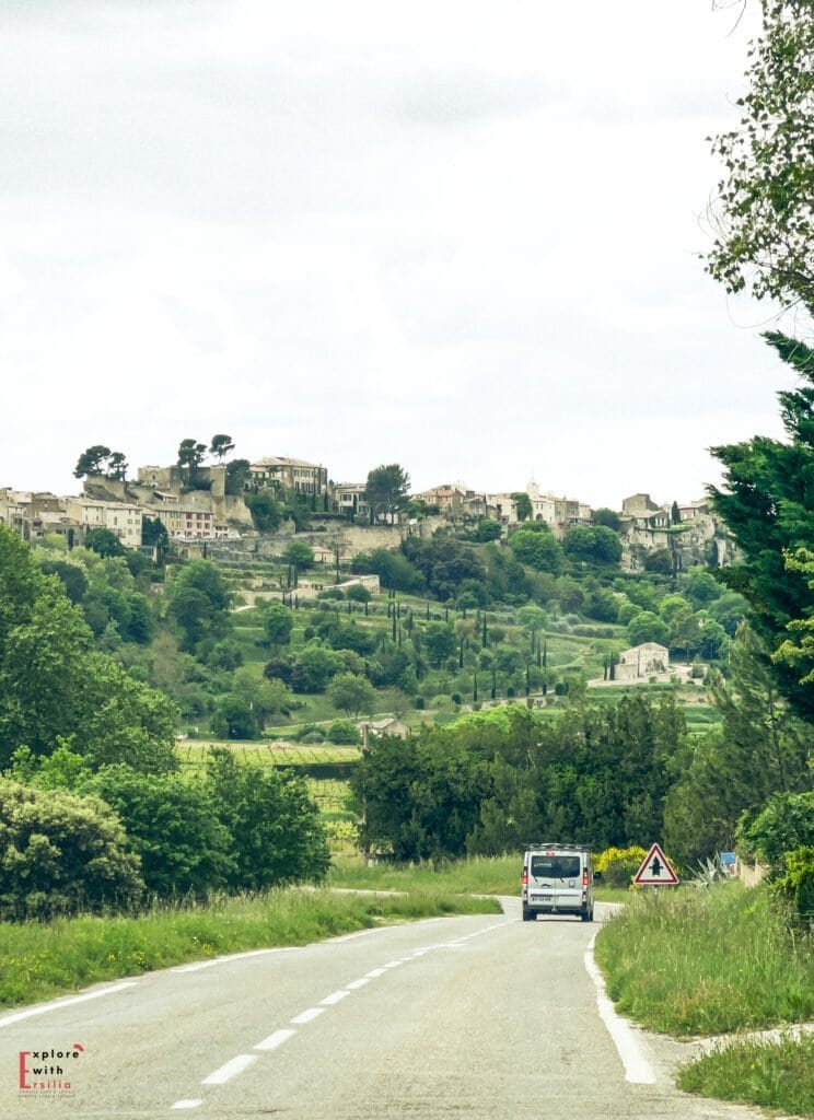 Driving through the scenic countryside of Provence toward the hilltop village of Lacoste, with a winding road framed by vineyards, cypress trees, and medieval stone homes—an unforgettable view on a day trip from Aix en Provence to one of the best villages to see in Provence.