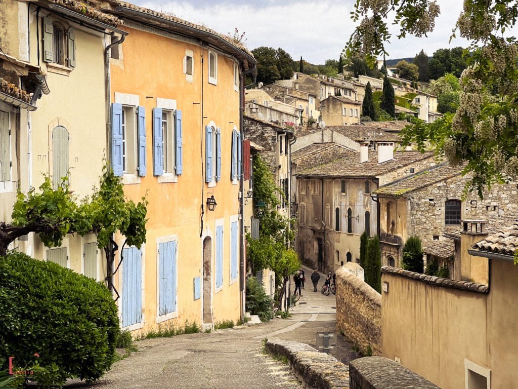 Charming street in Ménerbes, one of the best villages to see in Provence, with pastel-hued buildings, blue shutters, and cobbled lanes—an ideal stop on a day trip from Aix en Provence through the Luberon countryside.