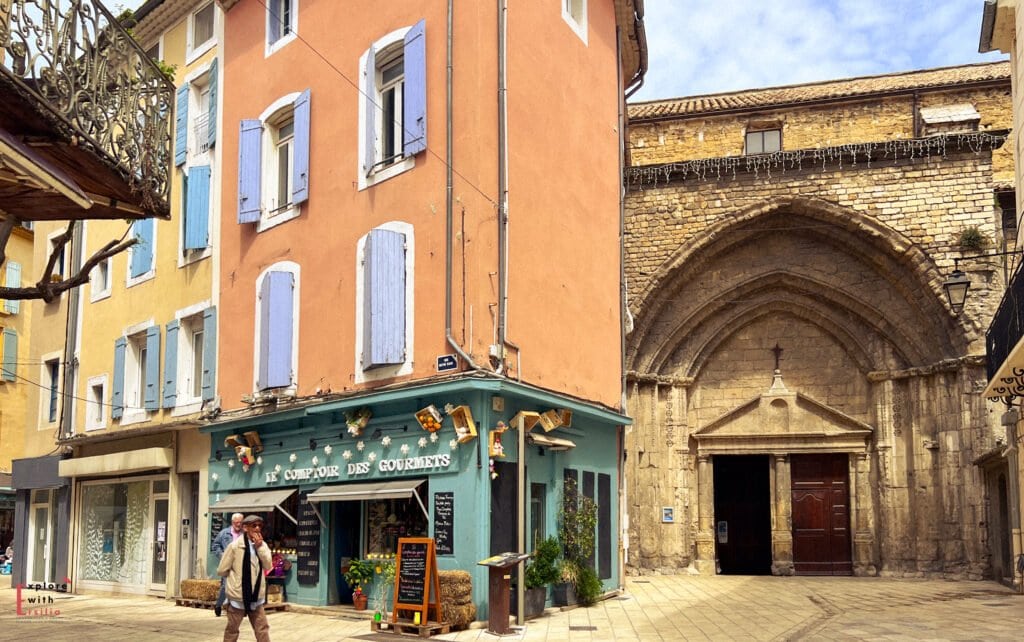 Colorful street scene in Orange, Provence, featuring pastel buildings with blue shutters, a quaint shop called “Le Comptoir des Gourmets,” and a medieval stone church with a grand arched entrance. A pedestrian strolls by under a partly sunny sky, adding life to this historic southern French town.