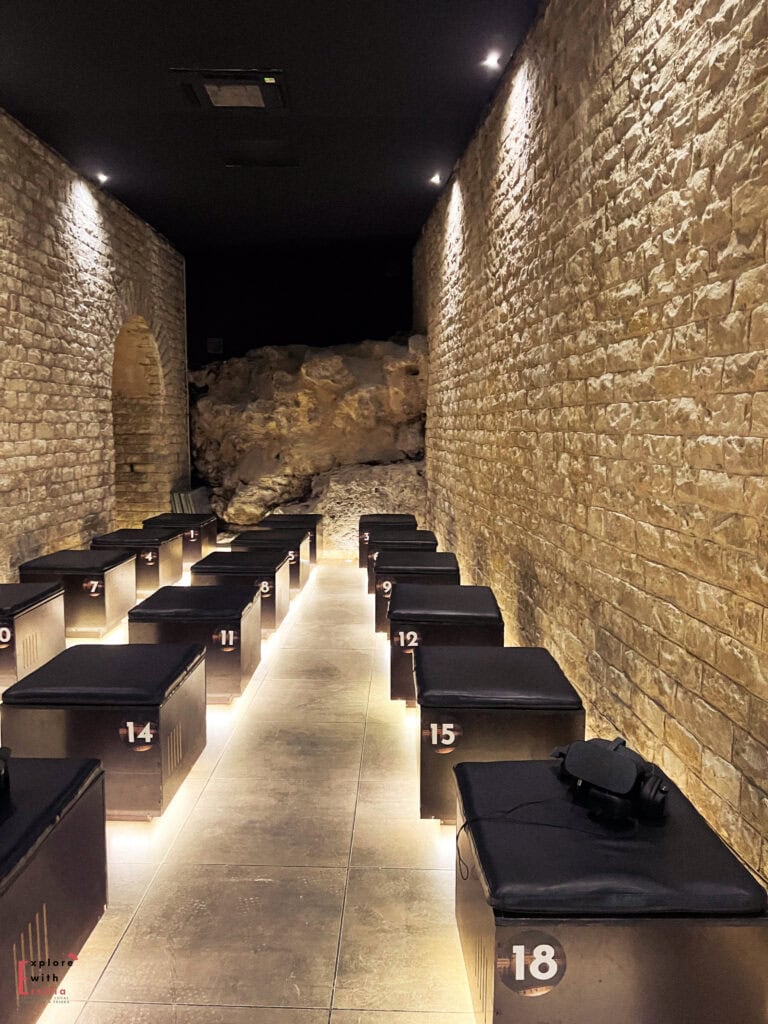 A narrow stone-walled room features two rows of black cushioned benches, each numbered and softly illuminated from below. At the end of the room, natural rock formations are visible, and a few benches have virtual reality headsets placed on them, suggesting an immersive exhibit experience.