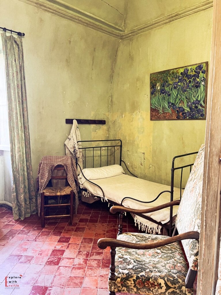 Interior of Van Gogh’s preserved bedroom at Saint-Paul de Mausole, featuring a simple iron-framed bed, wooden furniture, and reproduction paintings hanging on the aged, cream-colored walls.