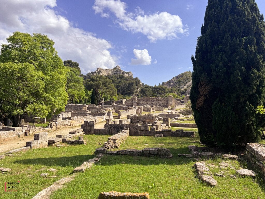 Ruins of the ancient Roman city of Glanum in Saint-Rémy-de-Provence, with scattered stone foundations, partial walls, and columns set against a backdrop of lush green trees and rocky hills under a partly cloudy sky. A tall cypress tree stands prominently to the right, framing the historical site.
