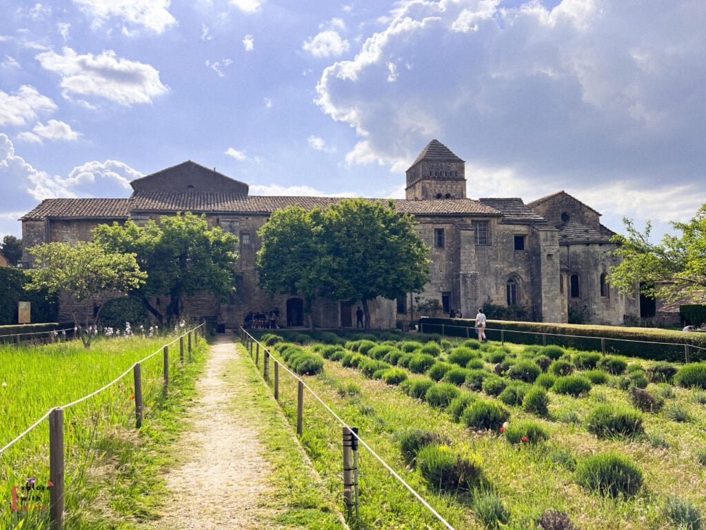 View of the historic Saint-Paul-de-Mausole monastery in Saint-Rémy-de-Provence, with a gravel path leading through lavender fields toward the stone building framed by leafy trees. Known for housing Vincent van Gogh during his stay, the serene site is bathed in soft sunlight under a dramatic, cloud-filled sky.