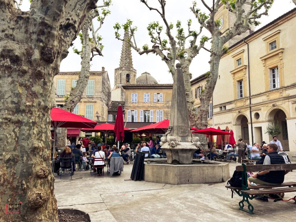 Bustling outdoor café scene in a plaza in Saint-Rémy-de-Provence, with red umbrellas, people dining at tables, and a central stone fountain surrounded by pruned plane trees. Historic buildings with pastel shutters and a church spire rise in the background, adding charm to the lively square.