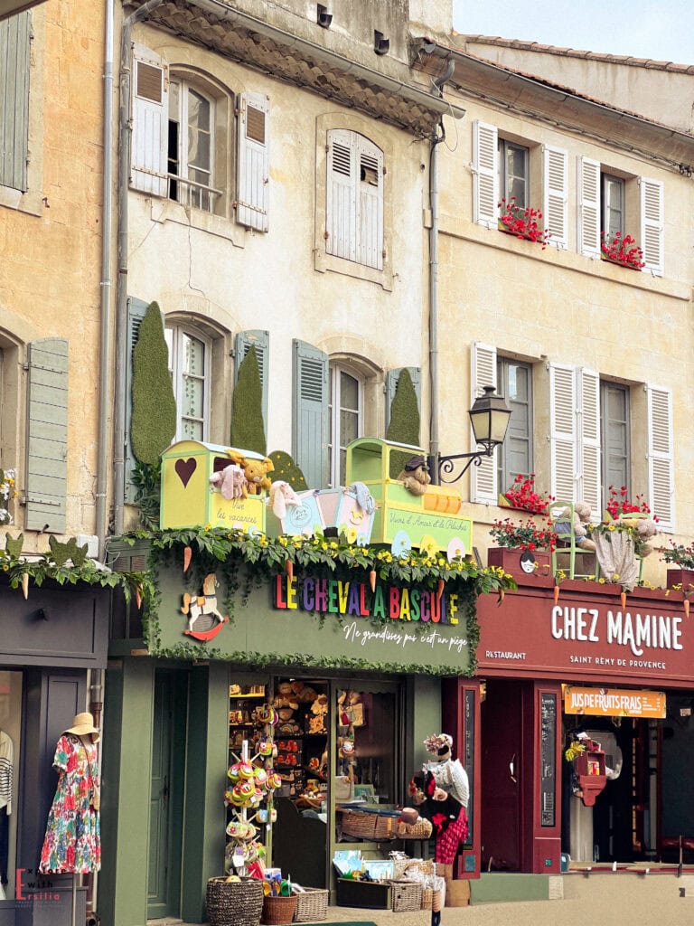 Colorful storefronts in Saint-Rémy-de-Provence, including the playful green façade of “Le Cheval à Bascule” toy shop decorated with plush animals, signs, and artificial greenery, and the adjacent red-fronted “Chez Mamine” restaurant. Above, classic shuttered windows and flower boxes with red blooms add to the charm of the historic stone buildings.