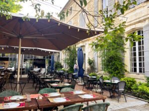 Charming outdoor restaurant in Saint-Rémy-de-Provence with neatly set tables under large umbrellas in a courtyard surrounded by ivy-covered stone walls and tall windows. The ambiance is cozy and inviting, perfect for a relaxed al fresco dining experience in a historic Provençal setting.