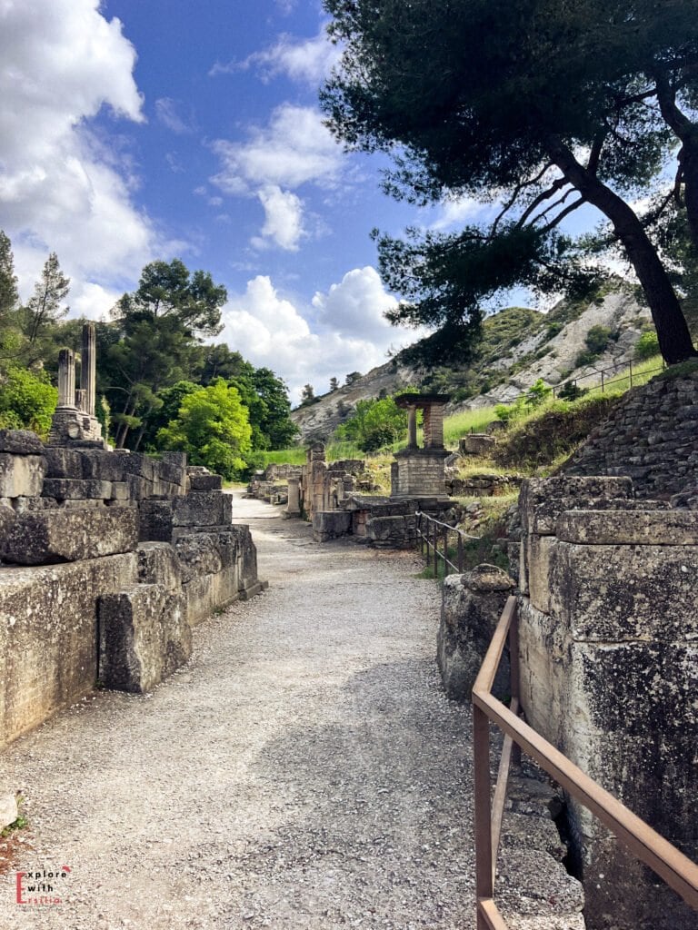 A stone pathway winds through the ancient Roman ruins of Glanum in Saint-Rémy-de-Provence, France, bordered by weathered stone blocks and partial columns. Surrounded by lush greenery and pine trees, the archaeological site sits beneath a rugged hillside under a partly cloudy sky.