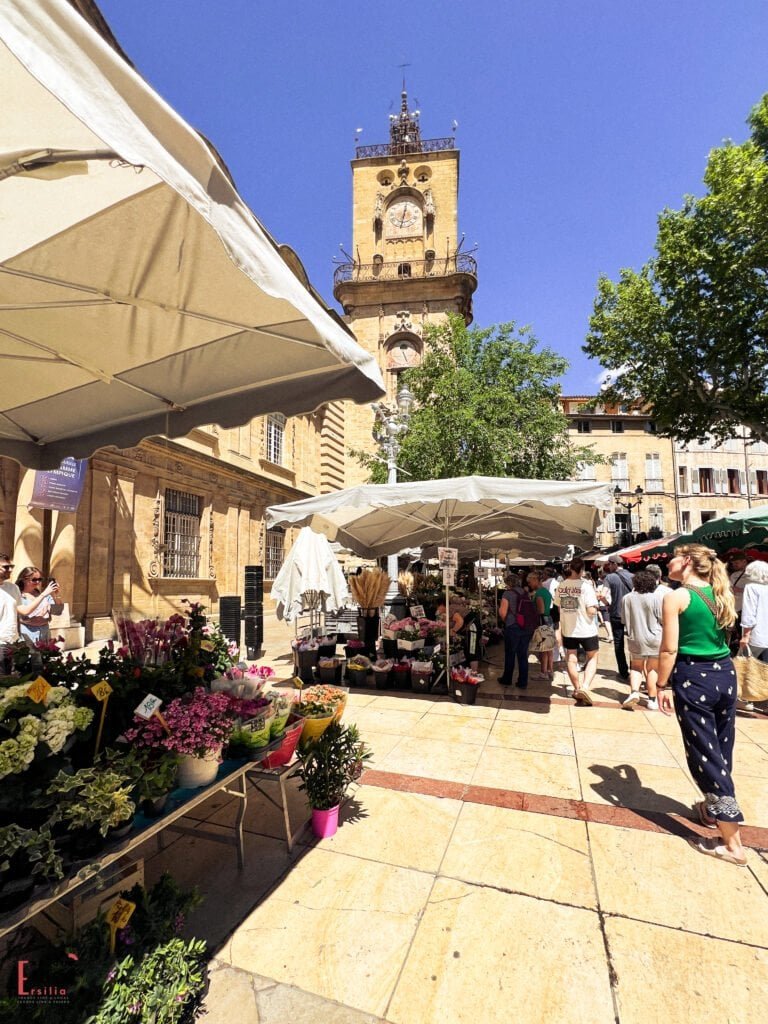 Outdoor flower market in Aix-en-Provence with colorful floral arrangements under white tents, bustling with shoppers on a sunny day. The iconic clock tower of Hôtel de Ville rises in the background against a clear blue sky, adding a historic charm to the lively square.