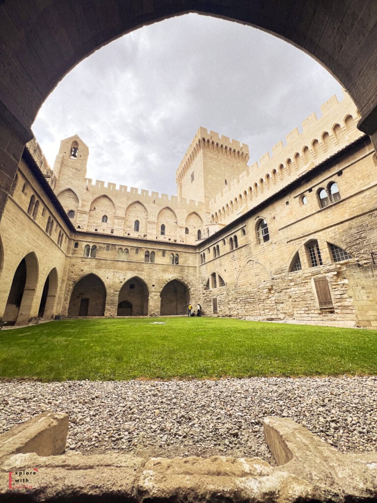 View of the inner courtyard of the Palais des Papes in Avignon, France, with arched stone walkways surrounding a grassy square. The towering medieval walls and battlements rise under a cloudy sky, showcasing the fortress-like architecture of the former papal palace.