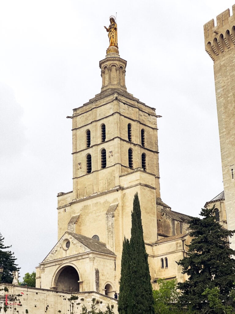 Close-up of the Avignon Cathedral, or Cathédrale Notre-Dame des Doms, with its tall Romanesque bell tower topped by a golden statue of the Virgin Mary. The historic stone façade stands against a cloudy sky, surrounded by greenery and medieval walls.