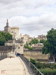 View of the historic Pont Saint-Bénézet, also known as the Pont d’Avignon, leading toward the fortified walls and towers of the Palais des Papes in Avignon, France. Visitors stroll along the stone bridge, with the golden statue of the Virgin Mary visible atop the cathedral in the background.