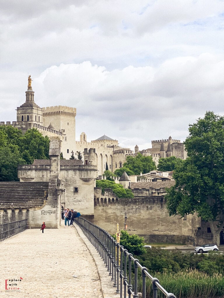 View of the historic Pont Saint-Bénézet, also known as the Pont d’Avignon, leading toward the fortified walls and towers of the Palais des Papes in Avignon, France. Visitors stroll along the stone bridge, with the golden statue of the Virgin Mary visible atop the cathedral in the background.