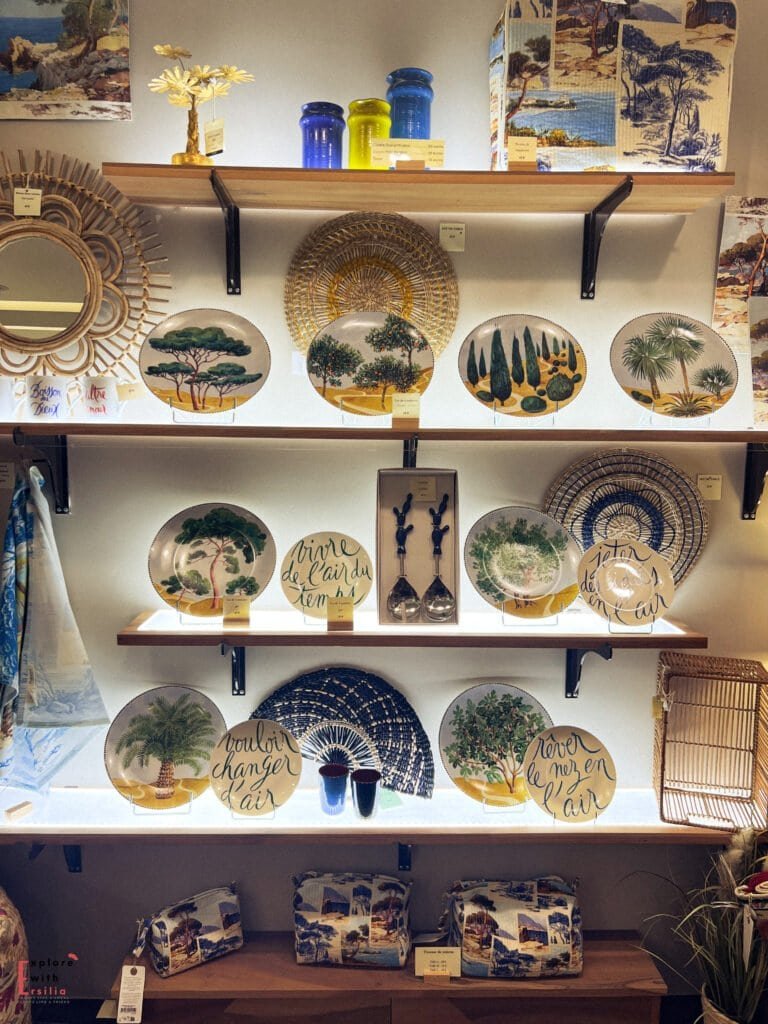 A display of decorative ceramic plates, fans, and Provençal-themed souvenirs in a shop in Avignon, France. The plates feature hand-painted trees, landscapes, and French phrases, capturing the charm of the region’s Mediterranean style.