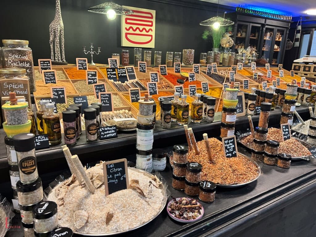 A colorful spice shop in Avignon, France, with neatly arranged jars and trays filled with herbs, salts, and spices labeled in French. The display features blends for raclette, vin chaud, and citrus-infused salts, creating a vibrant and aromatic scene.