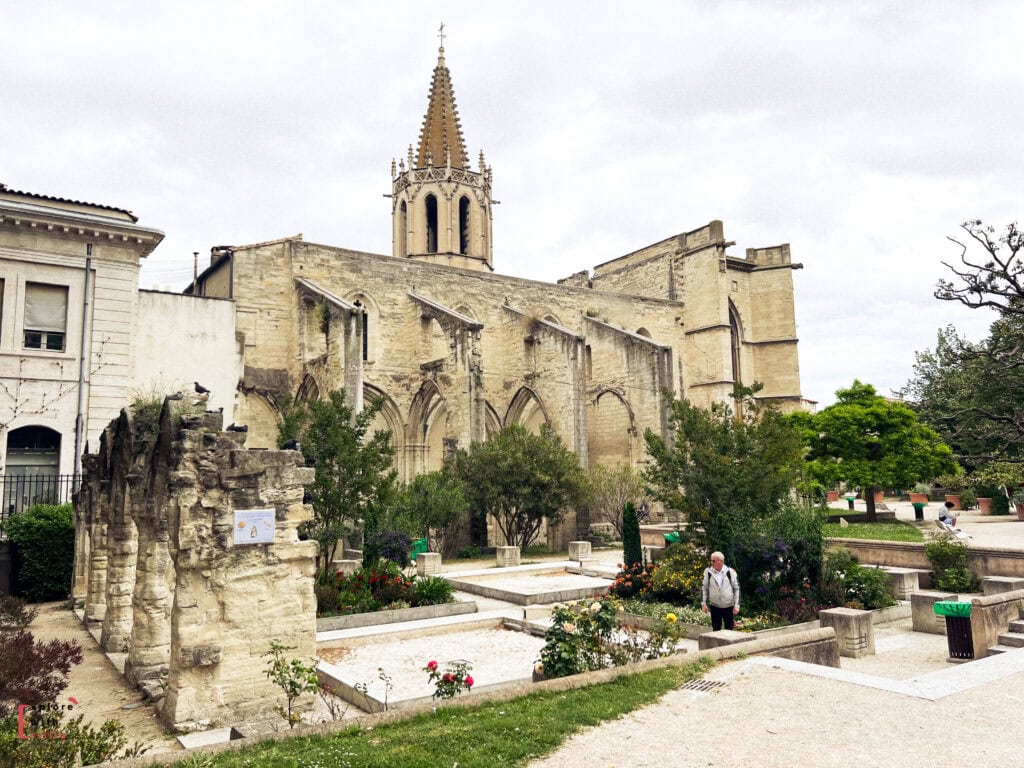 A peaceful garden with flower beds and stone ruins sits in front of a Gothic church in Avignon, France. The church’s pointed bell tower rises above the historic stone walls, while visitors stroll through the landscaped pathways.