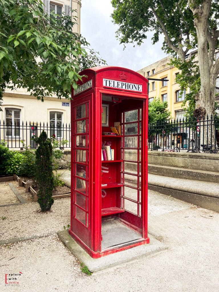 A classic red British telephone booth repurposed as a small public library stands in a charming square in Avignon, France. Books line the shelves inside, blending British nostalgia with a French community touch.