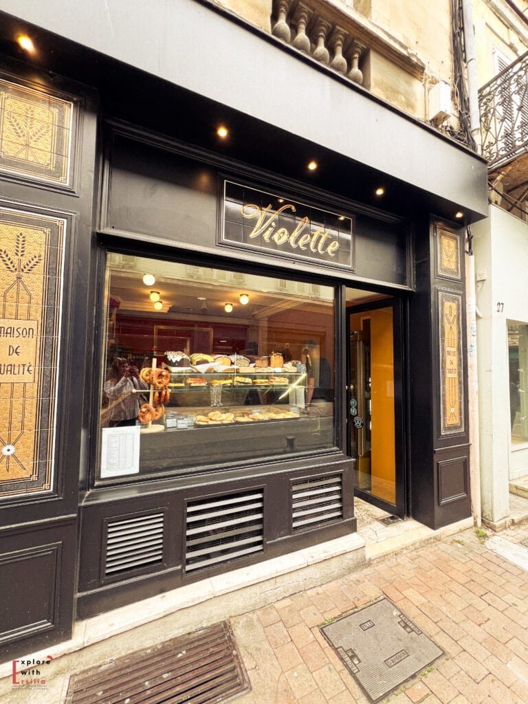 A charming bakery named "Violette" in Avignon, France, displays fresh pastries, breads, and pretzels in its window. The black storefront with gold lettering and decorative panels gives it an elegant, inviting Parisian-style look.