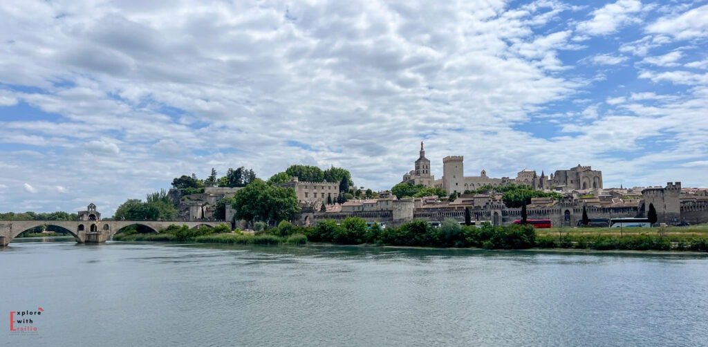 Panoramic view of Avignon, France, featuring the historic Saint-Bénézet Bridge (Pont d’Avignon) extending over the Rhône River and the imposing Palais des Papes rising above the medieval city walls. A dramatic sky of scattered clouds adds depth to the iconic skyline of this UNESCO World Heritage site.