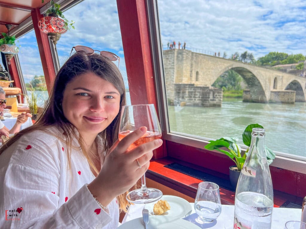 Woman enjoying a glass of rosé wine on a scenic boat ride in Avignon, with the famous Saint-Bénézet Bridge visible through the large windows beside her. The cozy interior features red wood framing, potted plants, and soft natural light reflecting off the Rhône River.