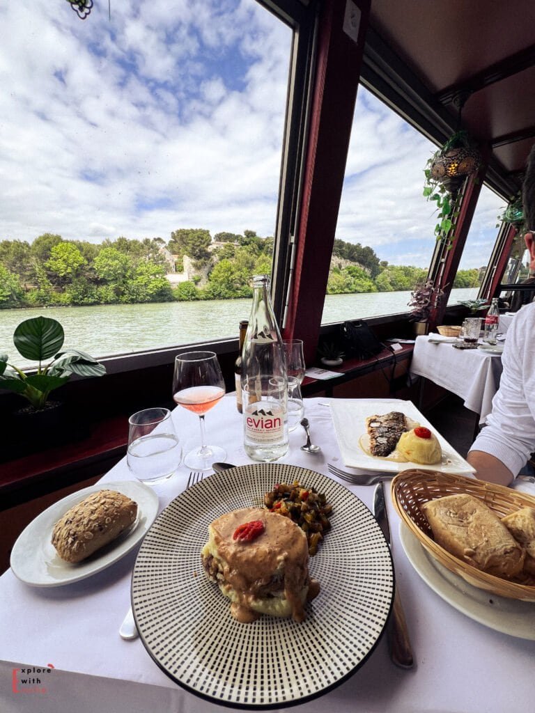 A beautifully plated French meal is served on a river cruise in Avignon, France, with views of the Rhône River through large windows. The table features a main dish topped with creamy sauce, ratatouille, fresh bread, a glass of rosé, and an Evian water bottle, creating a relaxed and scenic dining experience.