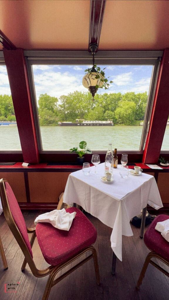 A cozy dining table set with empty glasses and coffee cups overlooks the Rhône River from a river cruise in Avignon, France. Large windows frame the scenic view of green trees and a passing boat, creating a peaceful post-meal moment.
