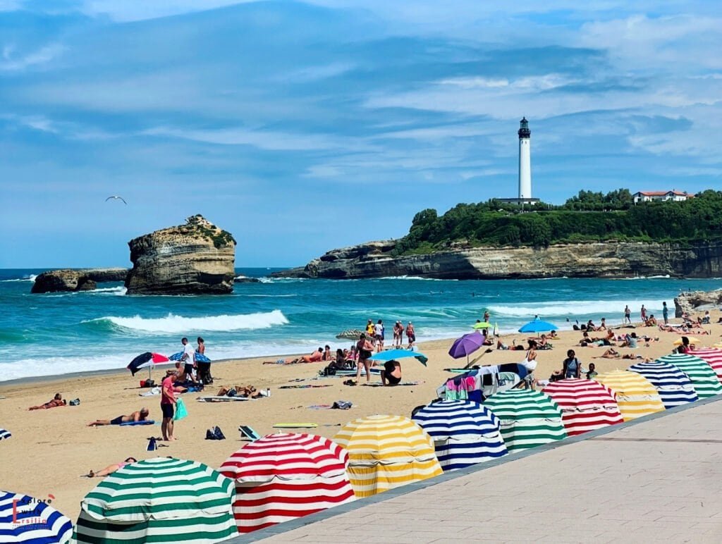 Vibrant beach scene in Biarritz featuring a row of colorful striped beach tents (traditional cabanas) in the foreground, with beachgoers enjoying the golden sand. In the background, the iconic Biarritz Lighthouse stands tall on a rocky headland, while a distinctive rock formation rises from the turquoise Atlantic waters. The beach is bustling with sunbathers and swimmers under a bright blue sky, capturing the quintessential French Basque coast summer atmosphere.