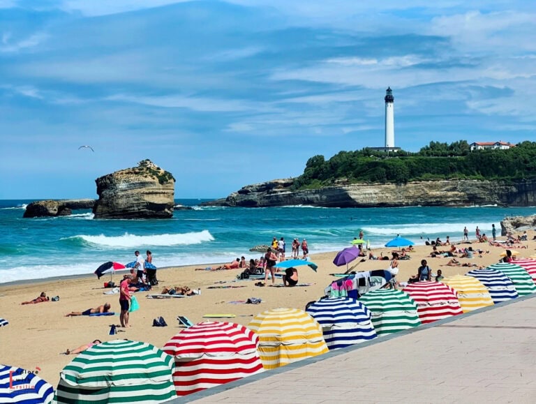 Vibrant beach scene in Biarritz featuring a row of colorful striped beach tents (traditional cabanas) in the foreground, with beachgoers enjoying the golden sand. In the background, the iconic Biarritz Lighthouse stands tall on a rocky headland, while a distinctive rock formation rises from the turquoise Atlantic waters. The beach is bustling with sunbathers and swimmers under a bright blue sky, capturing the quintessential French Basque coast summer atmosphere.
