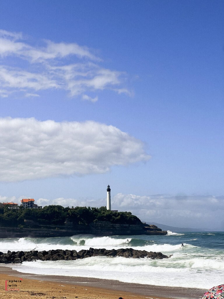 View of Biarritz Lighthouse (Phare de Biarritz) standing tall on a rocky cliff overlooking the Atlantic Ocean, as seen from Corsaires Beach. White waves crash against black rocks in the foreground, with surfers visible in the water. The iconic white lighthouse is surrounded by greenery and buildings under a brilliant blue sky with scattered clouds.