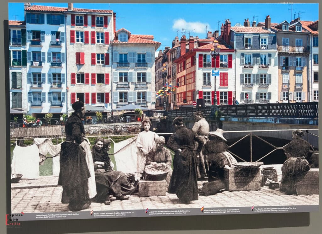 Composite historical image of Bayonne, France, showing traditional washerwomen from the early 20th century superimposed on a modern photograph of the city's colorful riverside buildings. The black and white historical photo shows women in long dresses washing clothes by the Nive River, while the contemporary background displays distinctive Basque architecture with white buildings featuring red and blue shutters. This creative juxtaposition illustrates the continuity between Bayonne's past and present along its iconic waterfront.