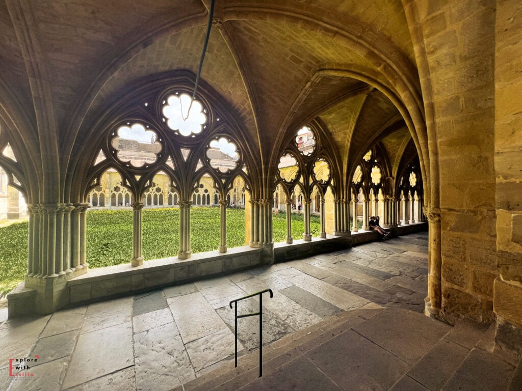 Interior view of a medieval Gothic cloister with elegant stone vaulted ceilings and quatrefoil-shaped arched openings overlooking a central courtyard with manicured greenery. The corridor features stone floor tiles and impressive ribbed vault architecture, with natural light filtering through the ornate tracery windows creating a serene, contemplative atmosphere.