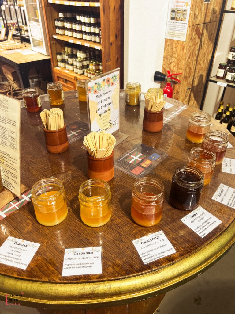 A circular wooden barrel table displaying an artisanal honey tasting setup at Loreztia Boutique. Small white ceramic dishes contain various honey varieties in different colors ranging from dark amber to clear, arranged in a circle with descriptive labels on wooden holders identifying each variety. Jars of honey for sale and wooden tasting sticks in terracotta holders are positioned in the center, with a second tasting barrel visible in the background of this rustic specialty food shop.